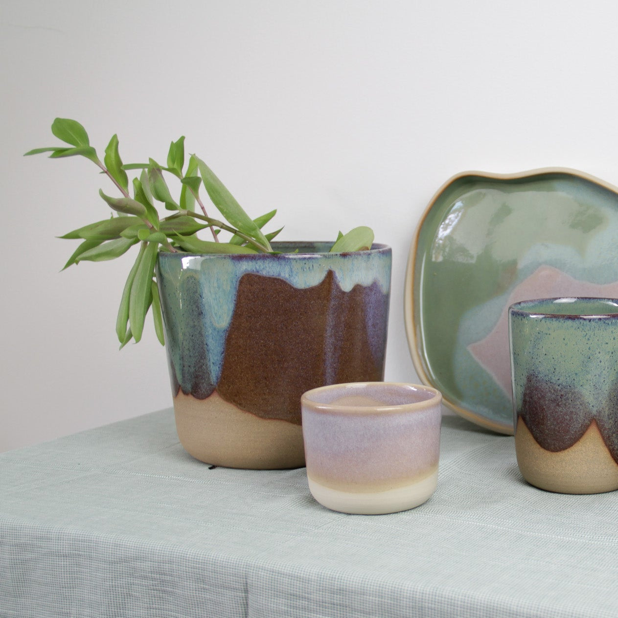 Photographie de poteries modernes et colorées. On voit un cache-pot avec une plante verte, des grands mugs en céramique et on aperçoit un plateau en céramique bicolore. Les céramiques présentent des nuances de verts et de roses.