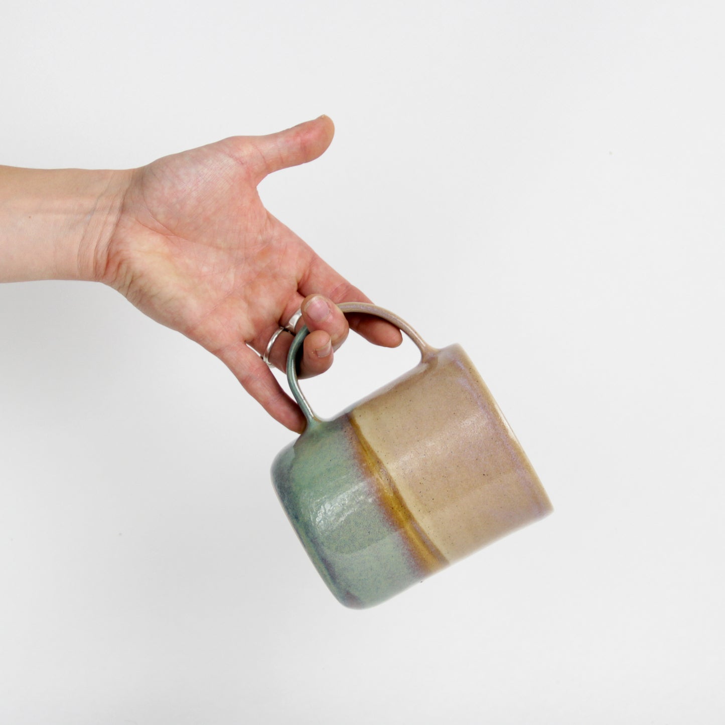 Image d'une main qui tient un grand mug par l'anse. Cette dernière est large et le mug est bicolore de la collection paysage vert.