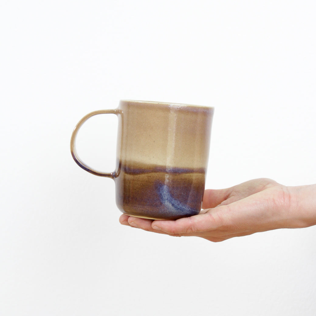 Image d'une main qui tient un grand mug avec une large anse. La tasse est un bicolore de la collection paysage violet