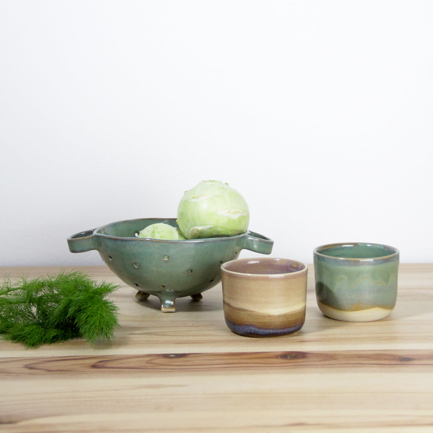 Image de plusieurs articles de vaisselle artisanale en céramique. On voit une passoire verte et deux petites tasses à café.