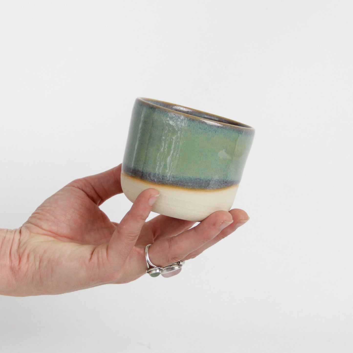 Image d'une main qui tient une petite tasse verte en céramique. La tasse est émaillée que sur sa partie haute et on peut voir la terre.