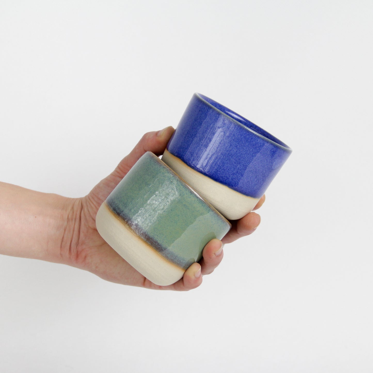 Image d'une main qui tient deux petites tasses en céramique, l'une est bleue et l'autre est verte.