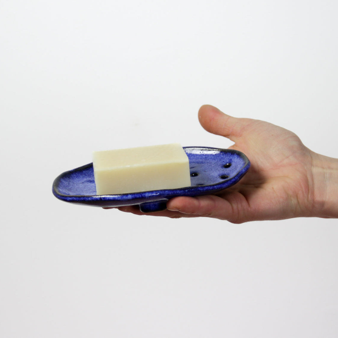Image d'une main qui tend un porte savon bleu foncé en céramique artisanale. Un savon est posé dessus.