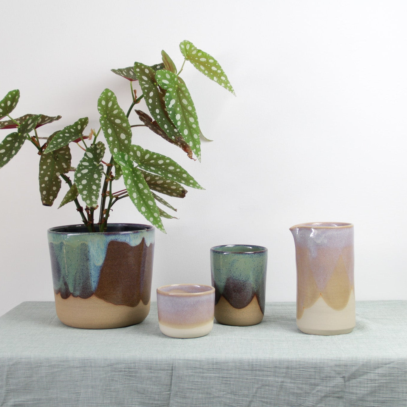 Image d'une table ou est posé dessus un cache-pot en céramique de couleur vert. Le pot contient une plante. À côté, il y a des tasses et un pichet rose.