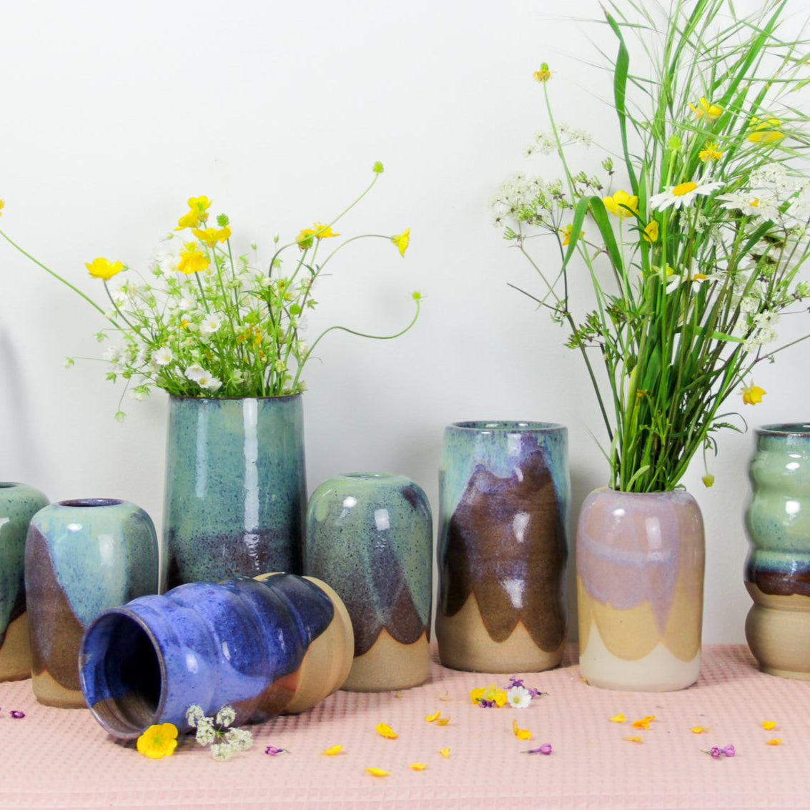 Photographie d'un ensemble de vases en céramique colorée. Il y a des soliflores, des grands vases, des plus petits. Certains vases contiennent des fleurs. Plusieurs couleurs : rose, vert, bleu.