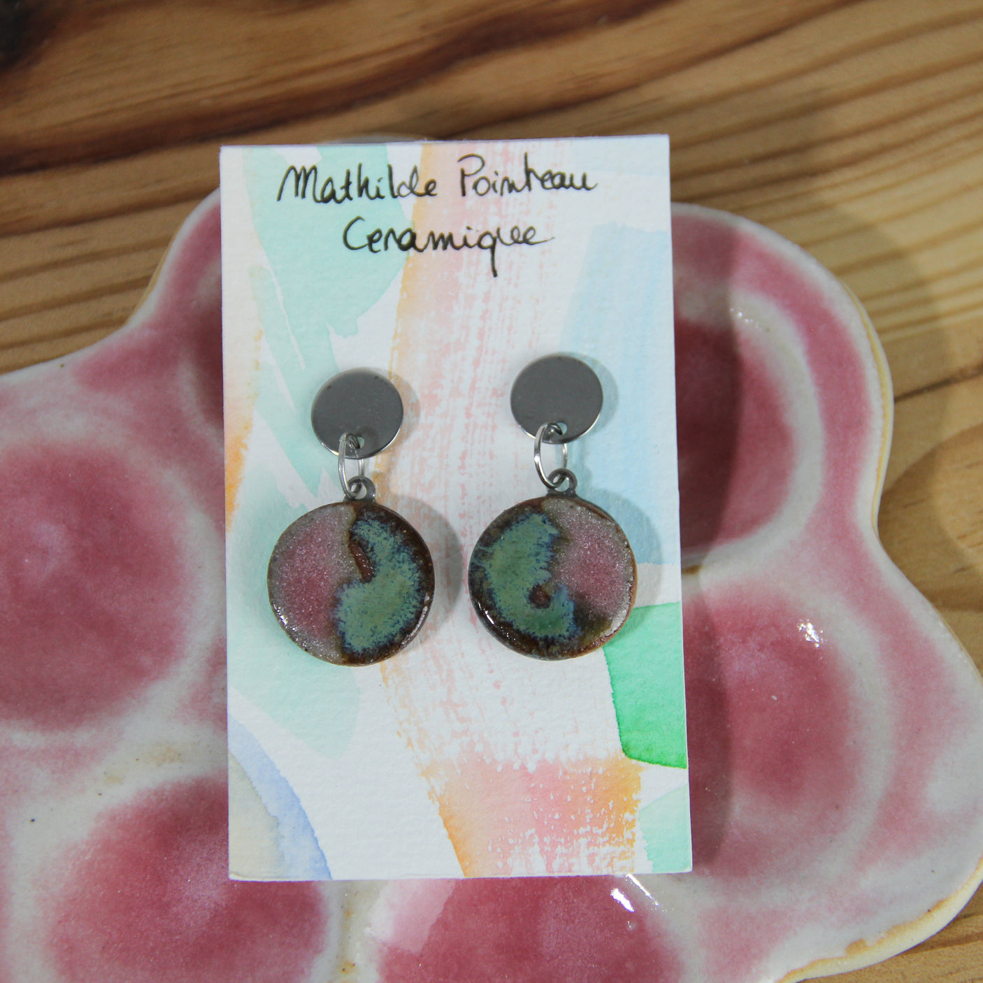 Image d'une paire de boucles d'oreilles en céramique rose et verte de forme ronde. Les boucles sont pendantes avec une accroche en acier inoxydable. 