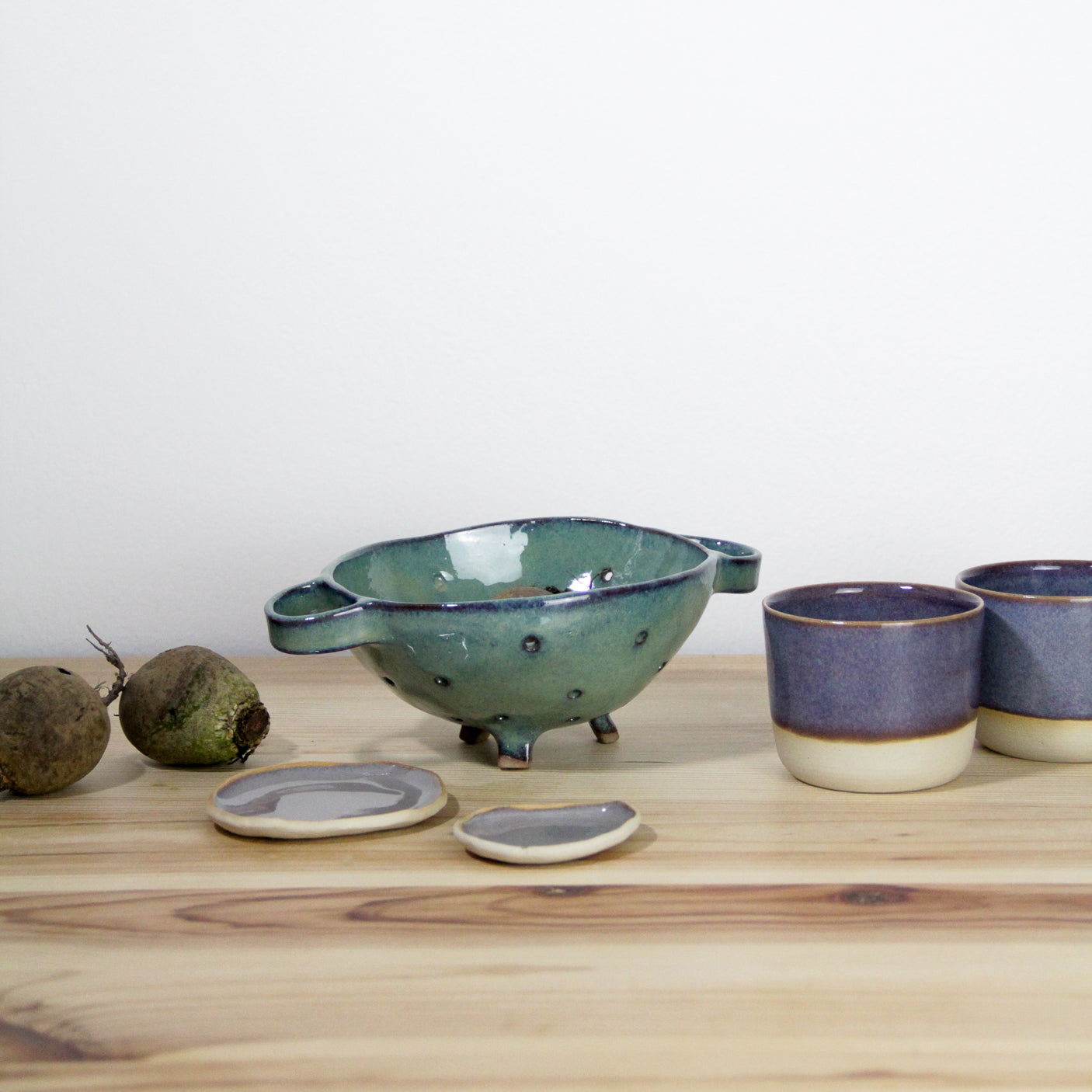 Image de plusieurs pièces de vaisselle en céramique artisanale. On voit une passoire verte faite main et deux petites tasses violettes.