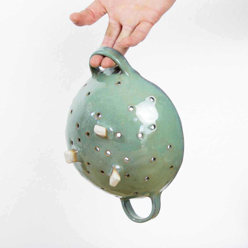 Image d'une passoire verte en céramique artisanale, une main la suspend et nous rends compte de la taille.