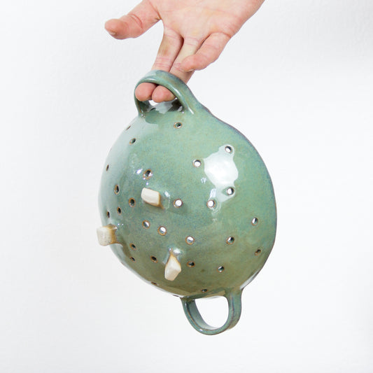 Image d'une passoire verte en céramique artisanale, une main la suspend et nous rends compte de la taille.