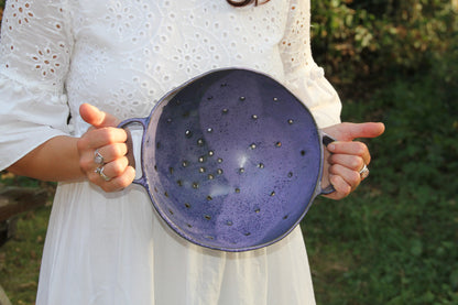 Image d'une femme qui tient une grande passoire en céramique artisanale. On peut voir l'échelle de la pièce. La passoire à deux grandes poignées de chaque côté.